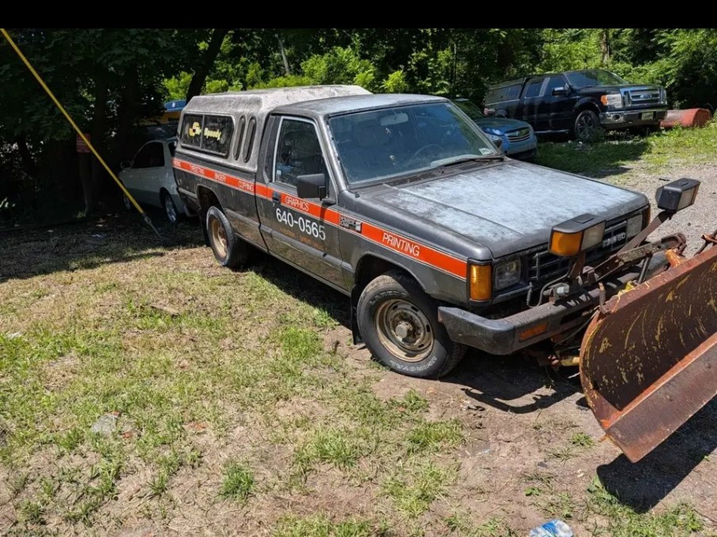Three-quarter view of the gray 1988 Dodge D50 with cap, plow, and work graphics